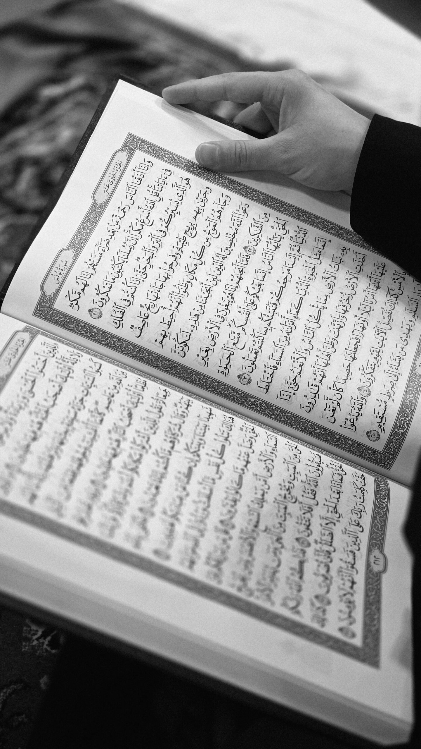 A person holding an open Quran, focusing on the intricate Arabic script in black and white.