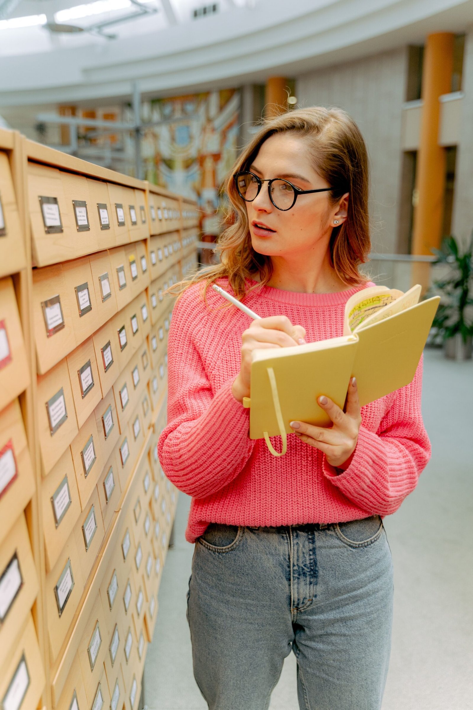 Young woman in glasses taking notes in library, wearing a pink sweater.